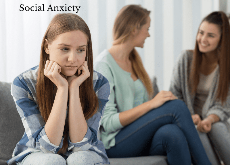 Young woman looking anxious and isolated while two others chat happily in the background, illustrating social anxiety in group settings.