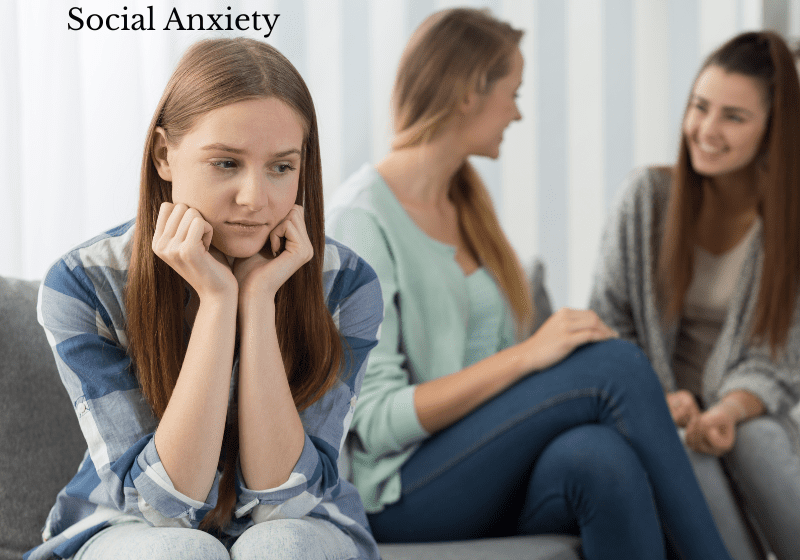 Young woman looking anxious and isolated while two others chat happily in the background, illustrating social anxiety in group settings.